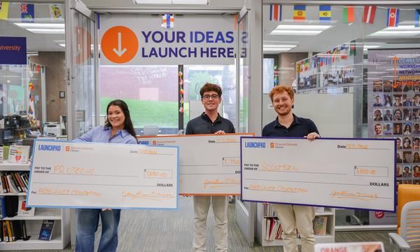three people holding large oversized checks in front of LaunchPad space
