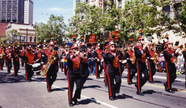 Archival image of marching band in red and black uniforms playing instruments in a parade with rainbow pride flags in background and on hats