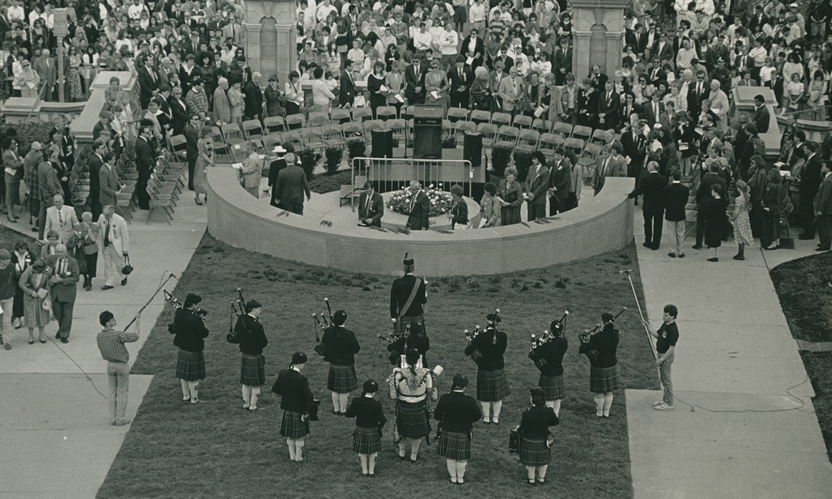 Black and white photograph of crowd gathered around a semi-circular wall. Several bagpipers standing behind the wall.