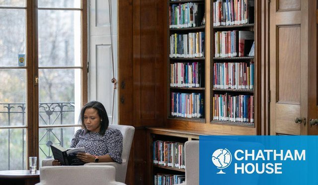 Black woman sitting next to a window and a large wooden book shelf in Chatham House reading a book with light blue and white logo for Chatham House