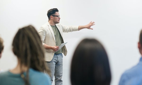 Teacher in a beige blazer in front of a room holding a notebook and gesturing at a white board