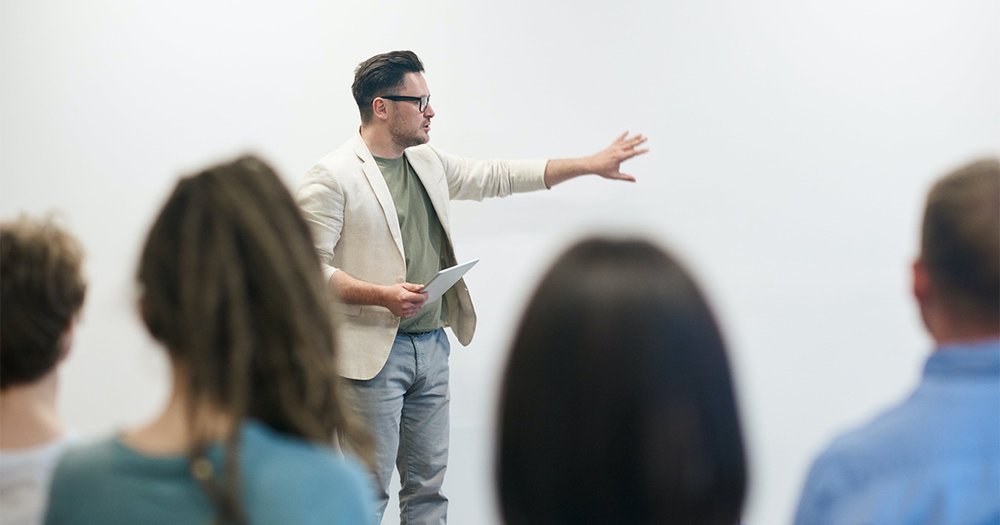 Teacher in a beige blazer in front of a room holding a notebook and gesturing at a white board