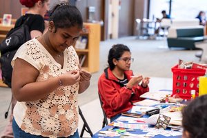 Two students working on crafts at a long table filled with paper products, scissors and stickers, with a red magazine bin nearby
