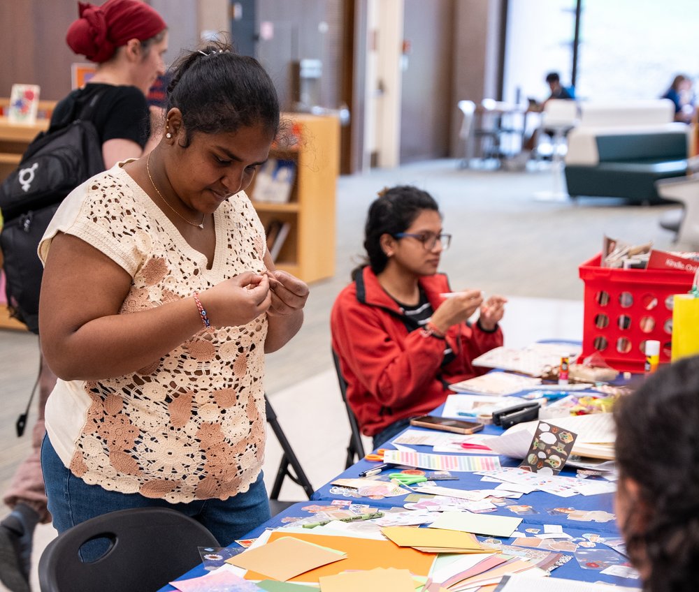Two students working on crafts at a long table filled with paper products, scissors and stickers, with a red magazine bin nearby