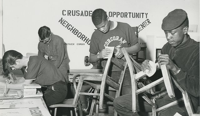 Black and white archival image of four people sanding wooden chairs and writing on a desktop with words "Crusade for Opportunity" on wall