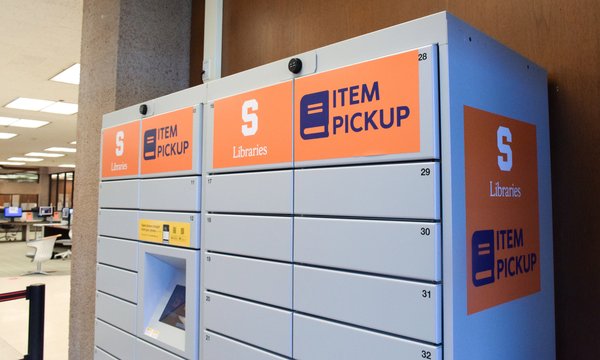 Gray lockers with orange signs that read "Item Pickup" and Syracuse University Libraries logo with touch screen in the middle