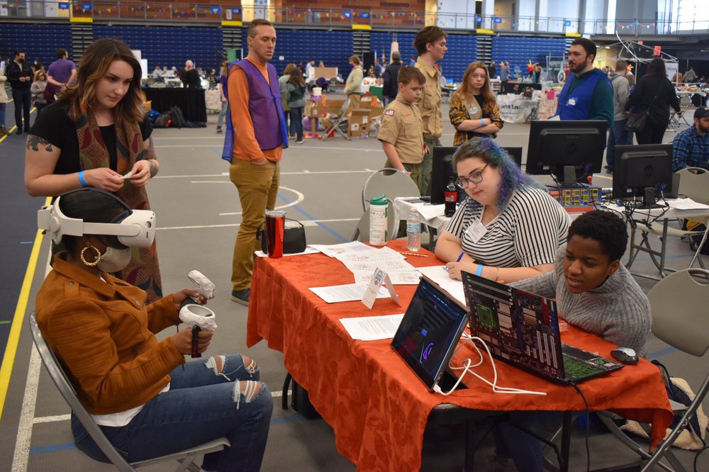 table with people sitting behind and in front of table, someone wearing virtual reality goggles and other people milling around in background