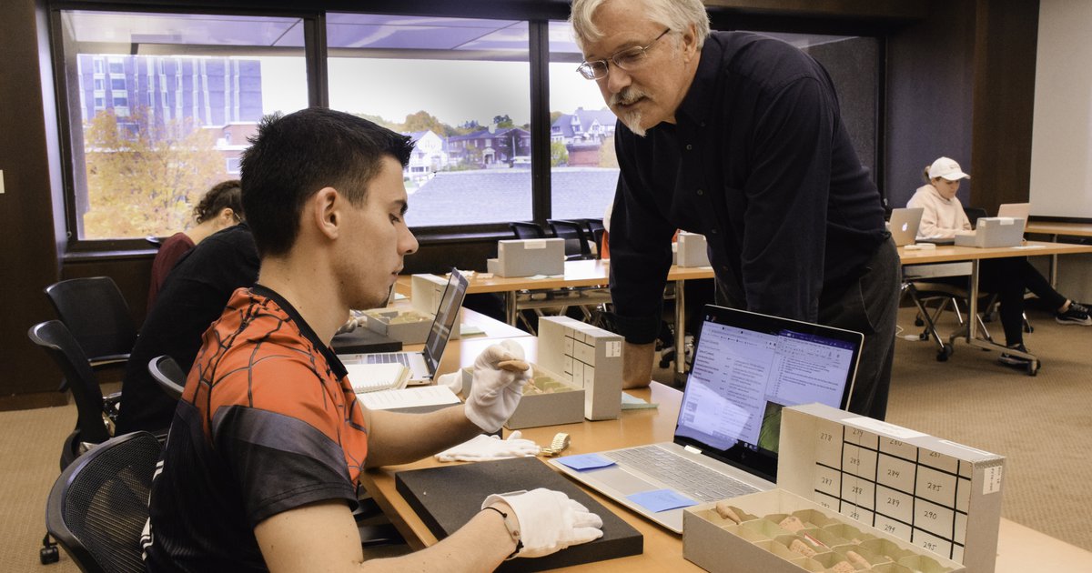 professor James Watts working with a student behind a table who's wearing white gloves and handling small cuneiform tablets from a box