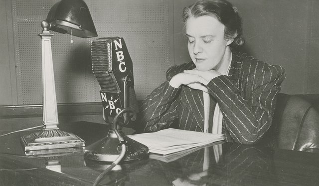 Black and white archival image of Dorothy Thompson sitting at a desk with papers, a lamp and a microphone labeled NBC