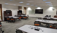 classroom with workstation tables and chairs around perimeter. Each set of table and chairs has monitor next to wall. Larger monitor in front of room