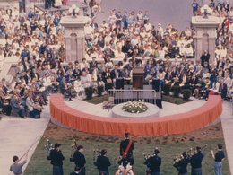 Color photograph of crowd gathered around a semi-circular wall covered in an orange cloth. Several bagpipers standing behind the wall.