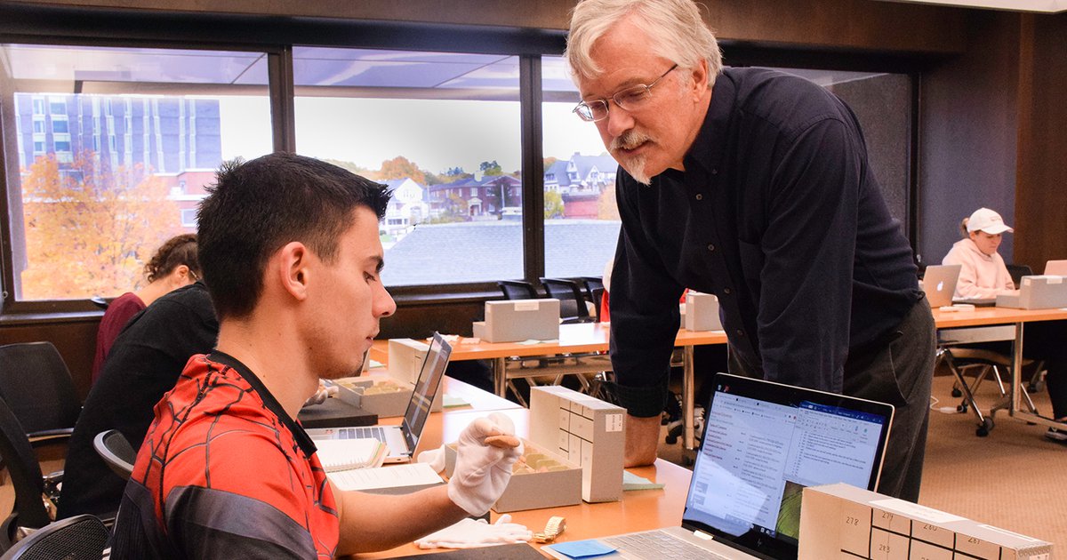 professor James Watts working with a student behind a table who's wearing white gloves and handling small cuneiform tablets from a box