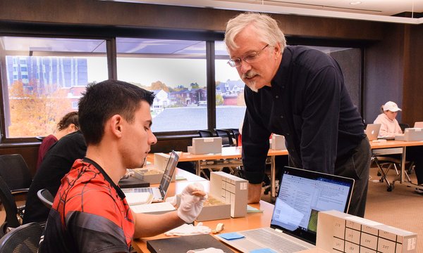 professor James Watts working with a student behind a table who's wearing white gloves and handling small cuneiform tablets from a box