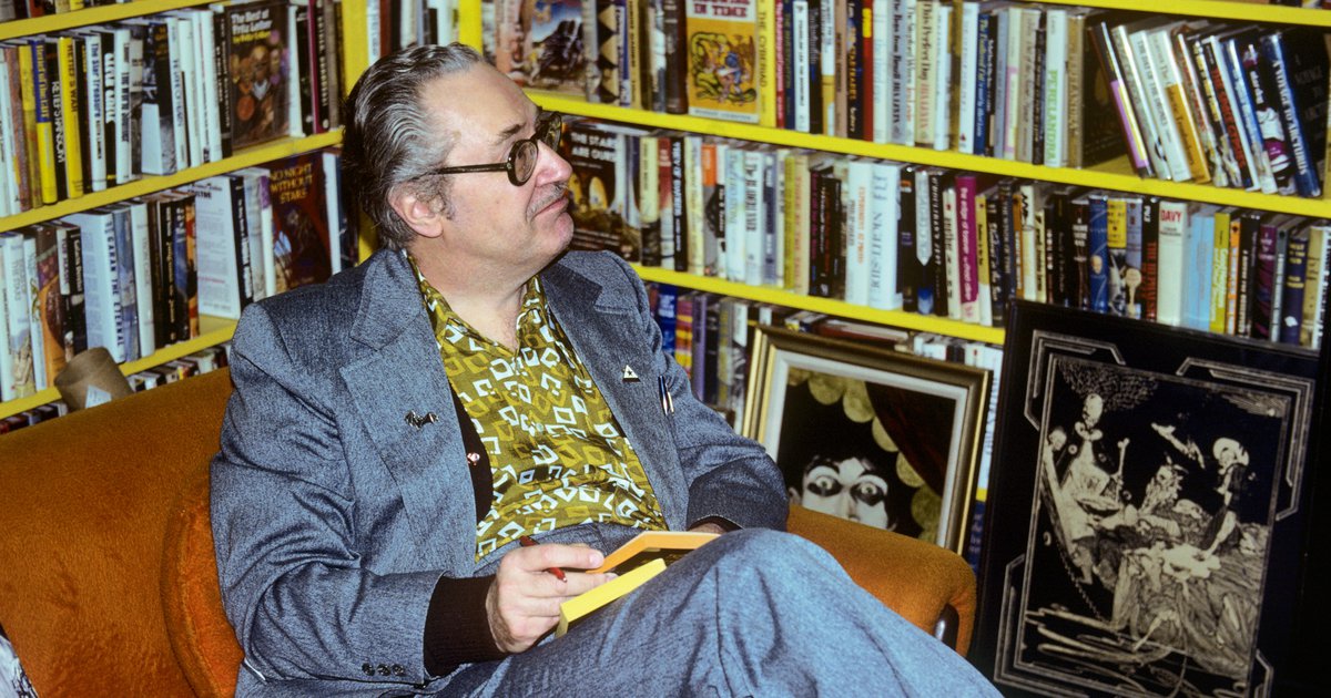 Forrest J Ackerman in his home, wearing a gray suit, yellow patterned shirt and glasses, sitting next to large, full, yellow book shelves