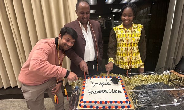 man wearing pink sweater bends down to cut cake that says "Congrats Founders Circle" while two other people standing behind table also appear to cut cake