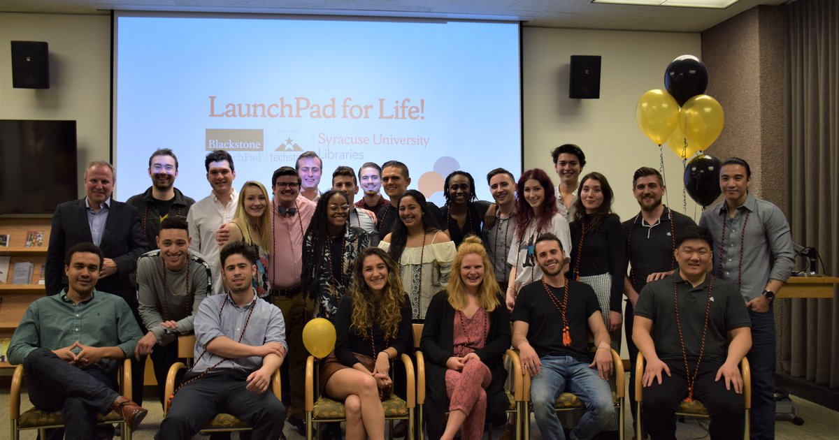 Group of 24 students posing in front of projector screen with black and yellow balloons on back right