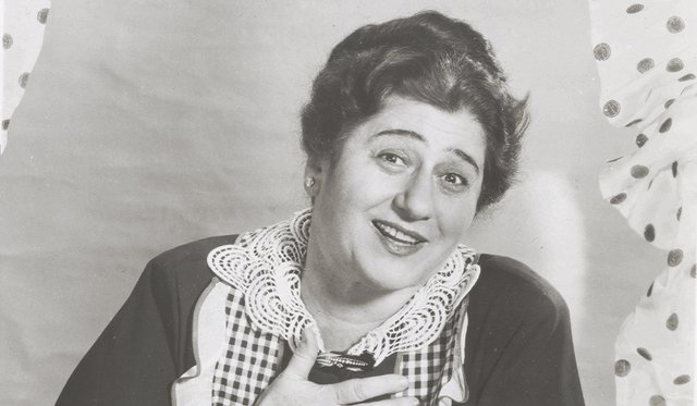 Black and white image of Gertrude Berg in a gingham apron smiling with hand on her chest