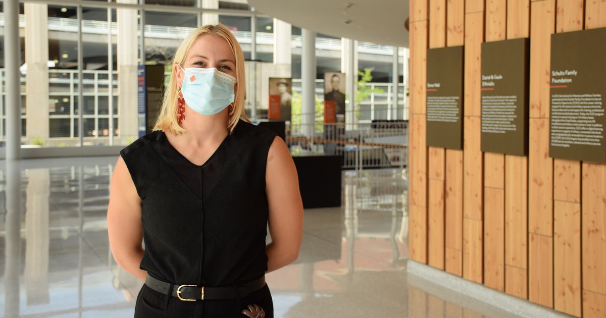 Grace Swinnerton wearing a mask and black tank top standing in the lobby of the Syracuse University Veterans Resource Center with wood slat wall