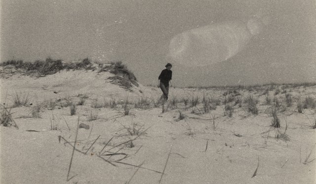 Black and white archival image of Grace Hartigan walking on a beach with sand dunes and wispy grasses