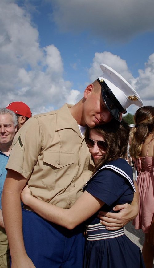 Man in uniform and a woman hugging with a crowd of people behind them.