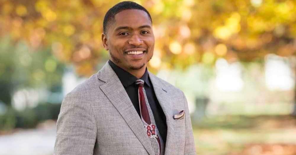 black man wearing suit standing outside with fall leaves on trees