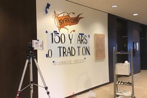 Exhibition wall that has Syracuse flag and beginning of letters that will read "150 Years of Tradition