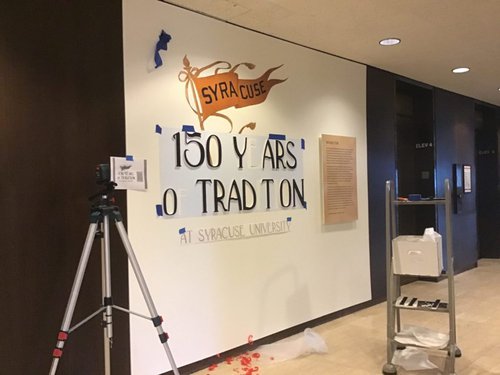 Exhibition wall that has Syracuse flag and beginning of letters that will read "150 Years of Tradition"
