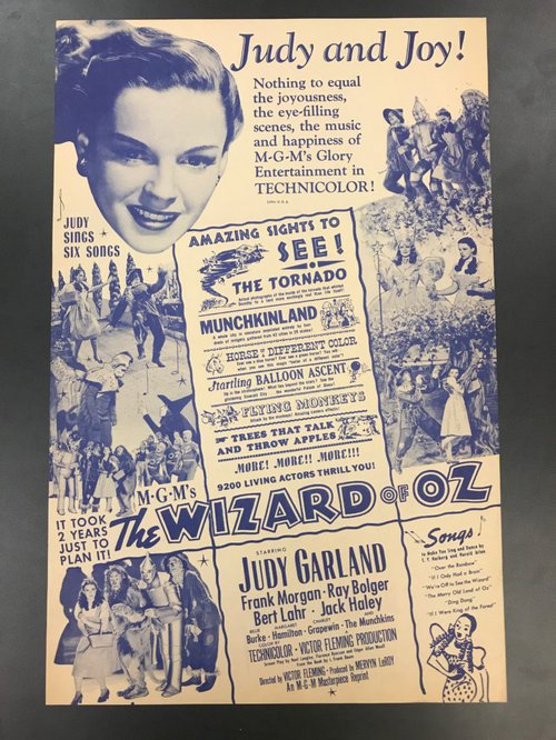 Blue and white poster advertising The Wizard of Oz film and picturing Judy Garland and multiple photographs depicting scenes from the movie.