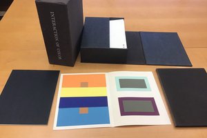 Paper with orange, yellow, dark blue, light blue, gray, and purple squares of paper attached inside, surrounded by black boxes and books on a wood table