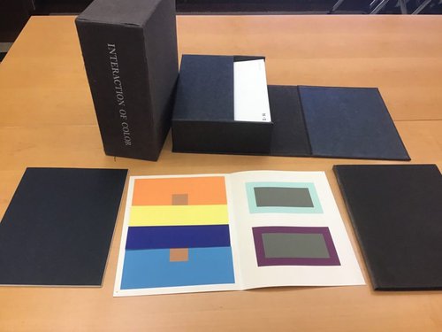 Paper with orange, yellow, dark blue, light blue, gray, and purple squares of paper attached inside, surrounded by black boxes and books on a wood table