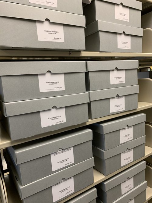 Fifteen blue boxes labeled Plastics Artifacts Collection on light colored metal shelves.