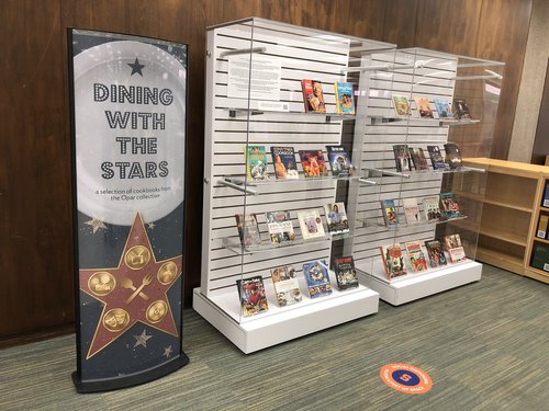 poster on left says "Dining with the Stars" written across plate and star with cutlery beneath, next to two glass display cases filled with cookbooks