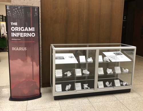 display poster in red reading "The Origami Inferno, Ikarus" next to glass display case with paper origami and descriptions
