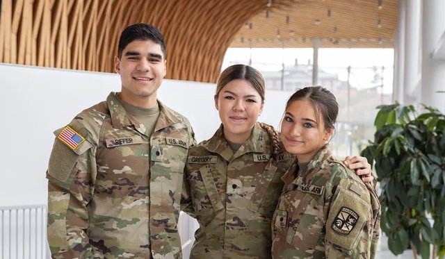Three students in camo military uniforms posing and smiling in front of curved architectural wooden slat ceiling