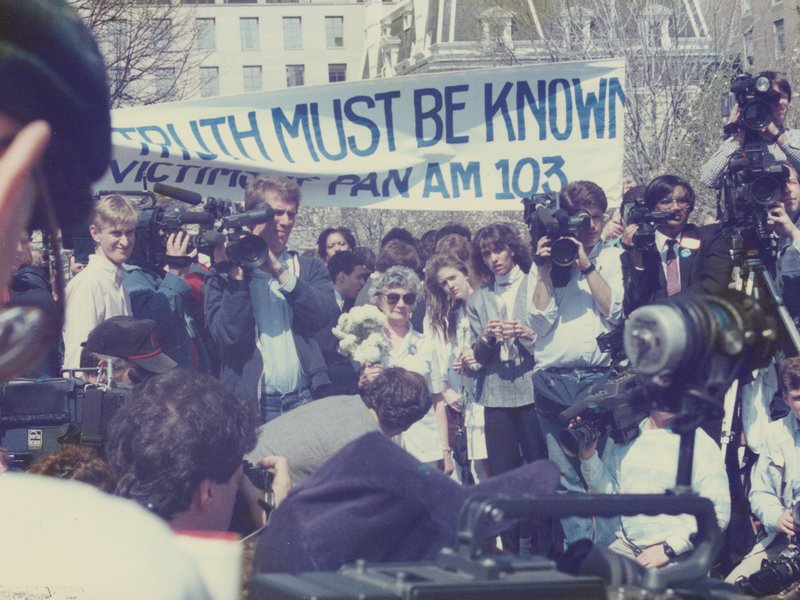 Group of people standing under a white banner with blue text that reads “Truth must be Known. Victims of Pan Am 103.” Surrounding the people at center are several camera crews.