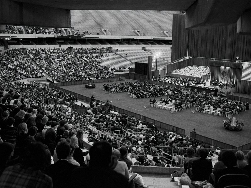 Black and white photograph of attendees seated in stadium for a memorial service. A stage with a speaker at a podium is visible on the right side.