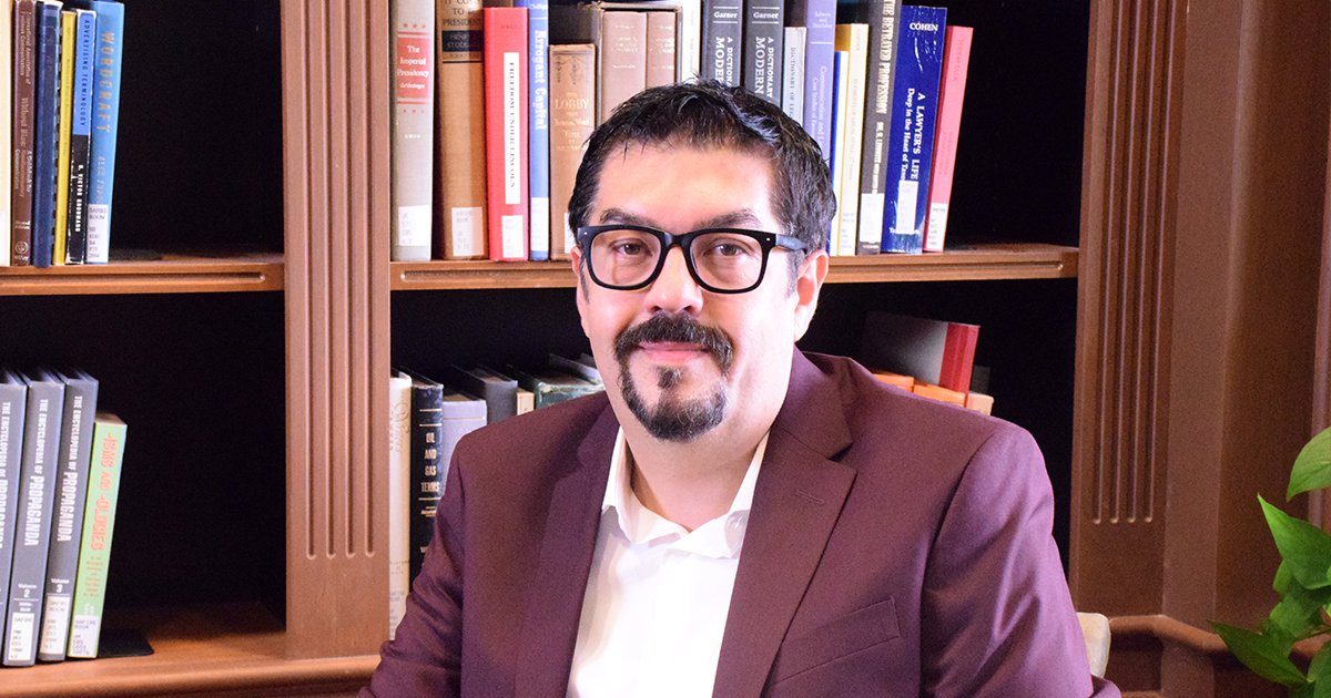 Juan Denzer in a maroon suit and glasses sitting in front of an ornate wooden bookshelf