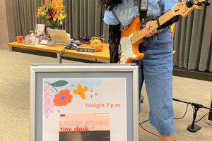 Lauren Juzang holding a guitar, standing in front the tiny desk sign