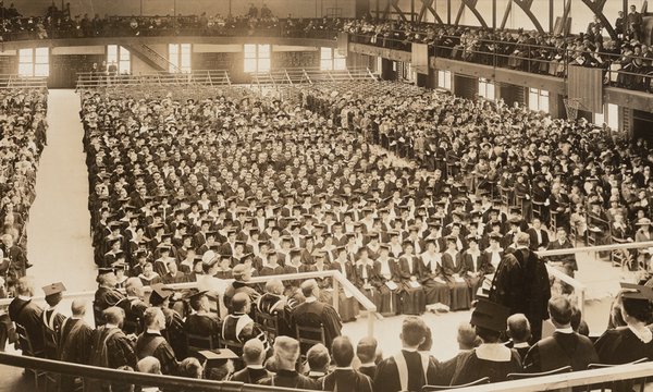 Sepia tone photograph of Chancellor James Roscoe Day speaking at Commencement ceremony in Archbold Gymnasium, 1916, with large crowd of graduates looking on. From the Syracuse University Photograph Collection, University Archives