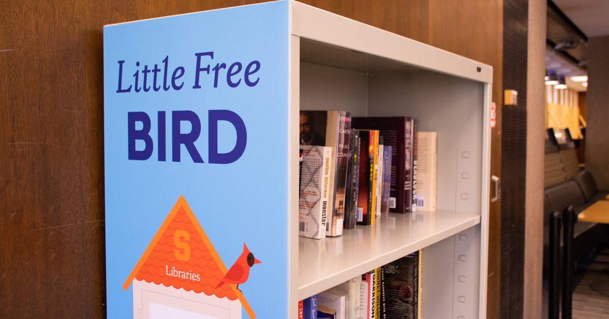 Gray metal bookshelf with two shelves of books with light blue sign on left that reads "Little Free Bird" in dark blue text with an orange Syracuse birdhouse and a cardinal