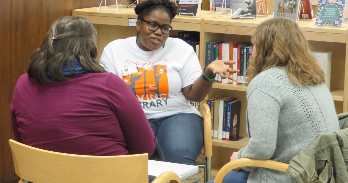 Three people sitting in chairs and talking in front of a book shelf