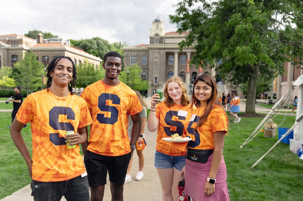 students standing in orange tie-dye t shirts with blue block "S" in front of grass on sidewalk by Carnegie Library