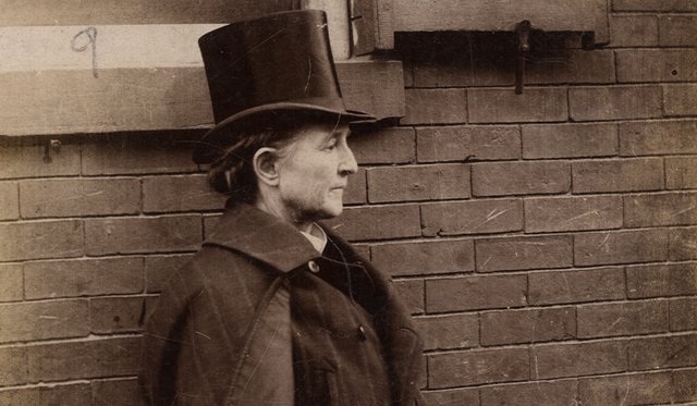 Sepia portrait of Mary Edwards Walker in profile wearing a tall black top hat and a mens' jacket, standing in front of a brick wall and window