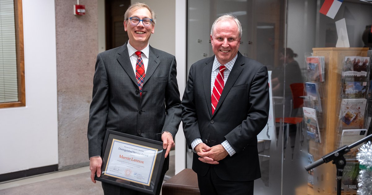 two men both wearing dark suits and red ties, the one on the left holding certificate