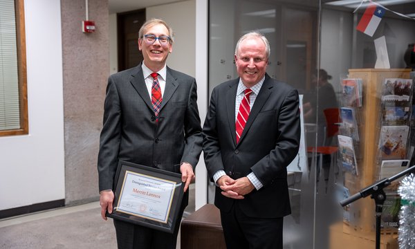 two men both wearing dark suits and red ties, the one on the left holding certificate