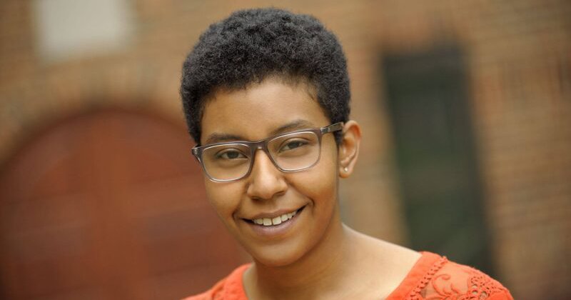 Monique Lassere standing in front of a brick building, wearing glasses and an orange shirt