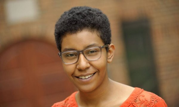 Monique Lassere standing in front of a brick building, wearing glasses and an orange shirt
