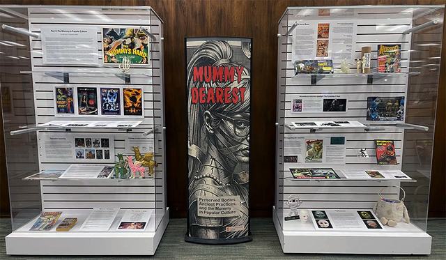 Two white background glass display cases with signage and artifacts about mummies in popular culture, with a center exhibit title banner that reads "Mummy Dearest" with an illustration of a woman with bandages and rough texture