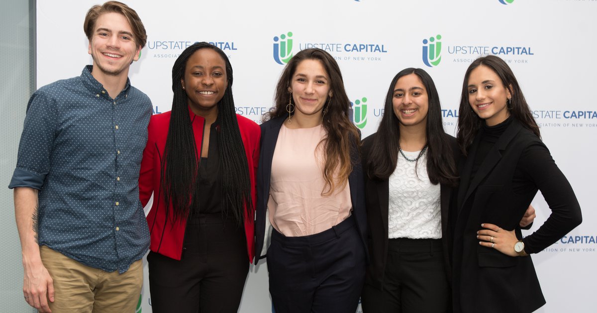 Five Syracuse University students wearing business casual clothes and standing in front of a white step and repeat banner with Upstate Capital logo