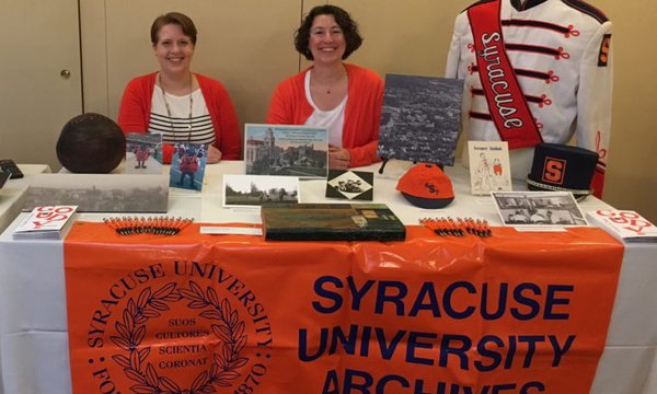 Two women wearing orange cardigans sitting behind Syracuse University Archives table, with memorabilia including band uniform on table
